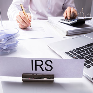 IRS Nameplate In Front Of Accountant Calculating Tax On Desk With Calculator And Laptop